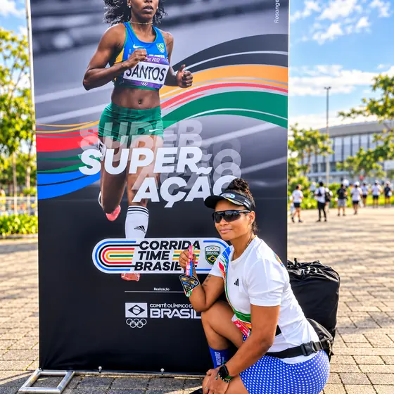 Patrícia agachada com a medalha diante do painel Superação da Corrida Time Brasil, do Comitê Olímpico do Brasil